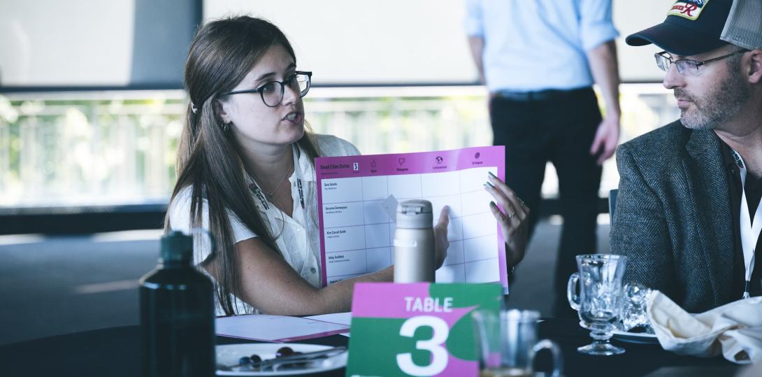 Female holding up a pink poster discussing it with a male sitting next to her.